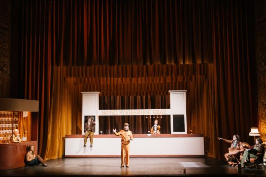 Foto mit gesamter Bühne, eine Person im Mittelpunkt vorne, zwei Personen sitzen flankierend auf der Bar dahinter