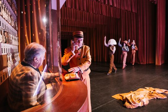 Foto mit 2 Ebenen, im Vordergrund 2 Personen bei der Portiersloge, eine telefoniert, im Hintergrund 3 Personen im Gleichschritt, jeweils einen Lampenschirm am Kopf