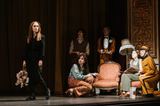 Foto mit sechs Personen auf der Bühne, eine Person im Mittelpunkt ins Publikum blickend, die anderen schauen sie an