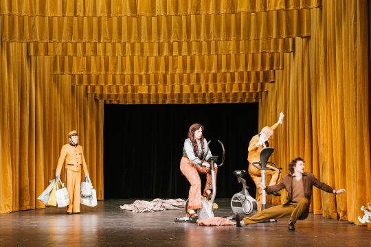 Foto mit 4 Personen auf der Bühne, 2 davon sitzen auf Ergometern, dunkelgelbe Vorhänge von der Decke und seitlich