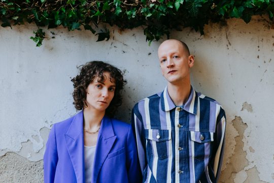 Foto mit Anna Klimovitskaya und Dominik Puhl an eine Wand gelehnt, in die Kamera blickend