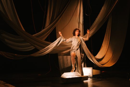 Foto mit Anna Klimovitskaya, sie ist in glänzend-beigefarbene, breite Stoffbänder eingehüllt, die von der Decke hängen