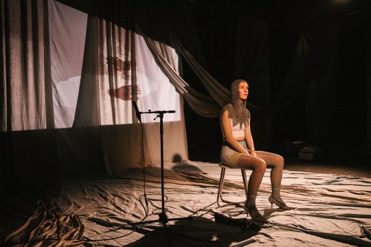 Foto mit Anna Klimovitskaya, mit einer Art Kettentuch über Kopf und Schultern gelegt, hinter ihr ist eine Leinwand mit Projektion zu sehen