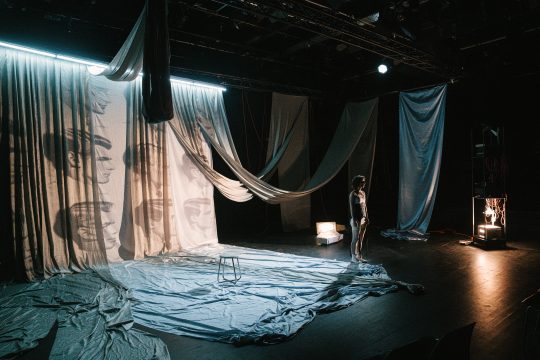 Foto mit Anna Klimovitskaya auf der Bühne, im Weitwinkel, man sieht Bänder von der Decke hängen und Tücher auf dem Boden liegen, eine Leinwand mit Männergesichtern dahinter