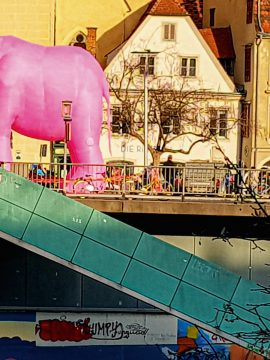 Foto mit einem aufgeblasenen rosa Nashorn oder Elefanten (Kopf ist nicht zu sehen) auf einer Brücke, Stiege hinunter in türkis und ein altes weißes Haus mit rotem Dach im Hintergrund