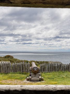 Foto mit einem Ausblick durch eine Öffnung aufs offene Meer, davor steht eine alte Kanone, bewölkter Himmel