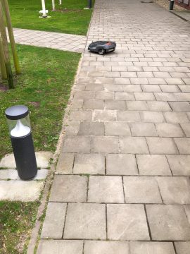 Foto mit einem gepflasterten Weg, darüber fährt ein Rasenroboter, links etwas Rasenfläche zu sehen, daneben eine Wegleuchte