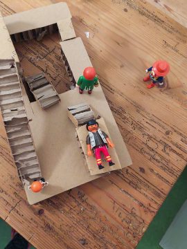 Foto mit Blick von oben auf einen Holztisch, darauf ein Stockwerk mit Stiege aus Karton und Playmobil-Spielfiguren