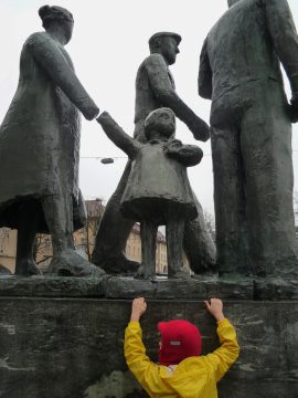 Foto mit Statuen auf einem hohen Sockel, 4 Figuren, darunter ein Kind zu sehen, ein kleines Kind mit gelber Jacke und roter Kappe versucht die Figuren auf dem Sockel zu erreichen