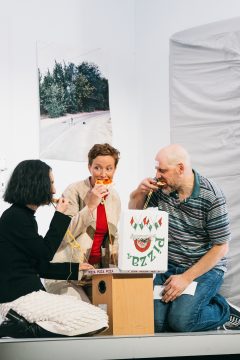Zwei Frauen, ein Mann hockend vor einem kleinen Tisch, sie essen Pizza