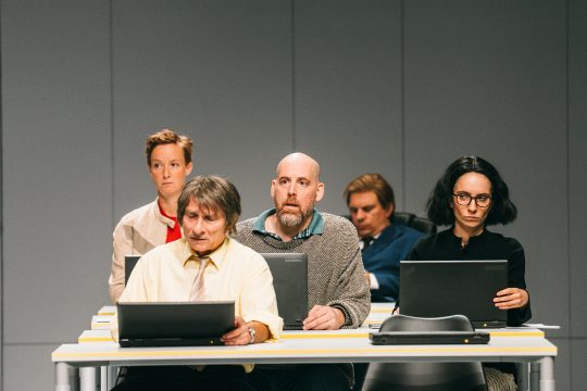 Fünf Personen sitzen in Reihen an Tischen, erinnert an Schulklasse