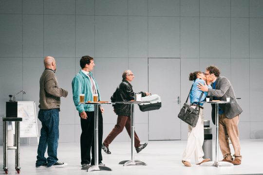 Fünf Personen auf einer Bühnen, zwei küssen sich, zwei hinter einem Stehtisch, eine Person läuft mit einer Tierbox durchs Bild