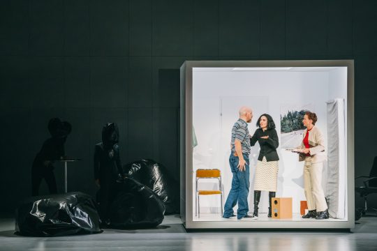 Ein Mann und zwei Frauen in einer hell erleuchteten Box, daneben zwei Personen im schwarzen Hintergrund